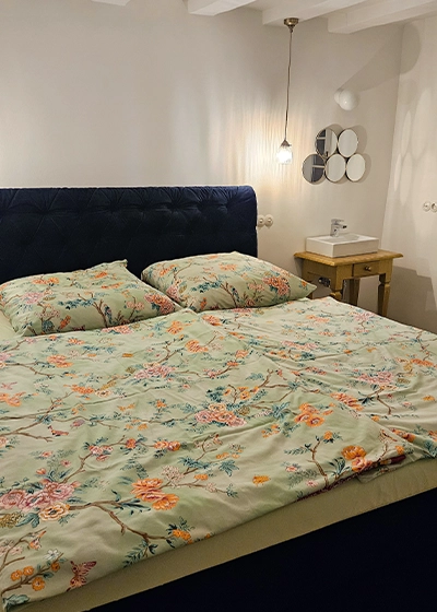 Schlafzimmer | Das Gloria Doppelbett bezogen mit einem grünen Bezug,welcher mit rosa und orangenen Blumen auf Ästen verziert ist. Rechts in der Ecke steht ein kleiner Waschtisch, mit einem Waschbecken. Darüber fünf kleine runde Spiegel. An der Decke hängt eine Glühbirne an einem langen Kabel